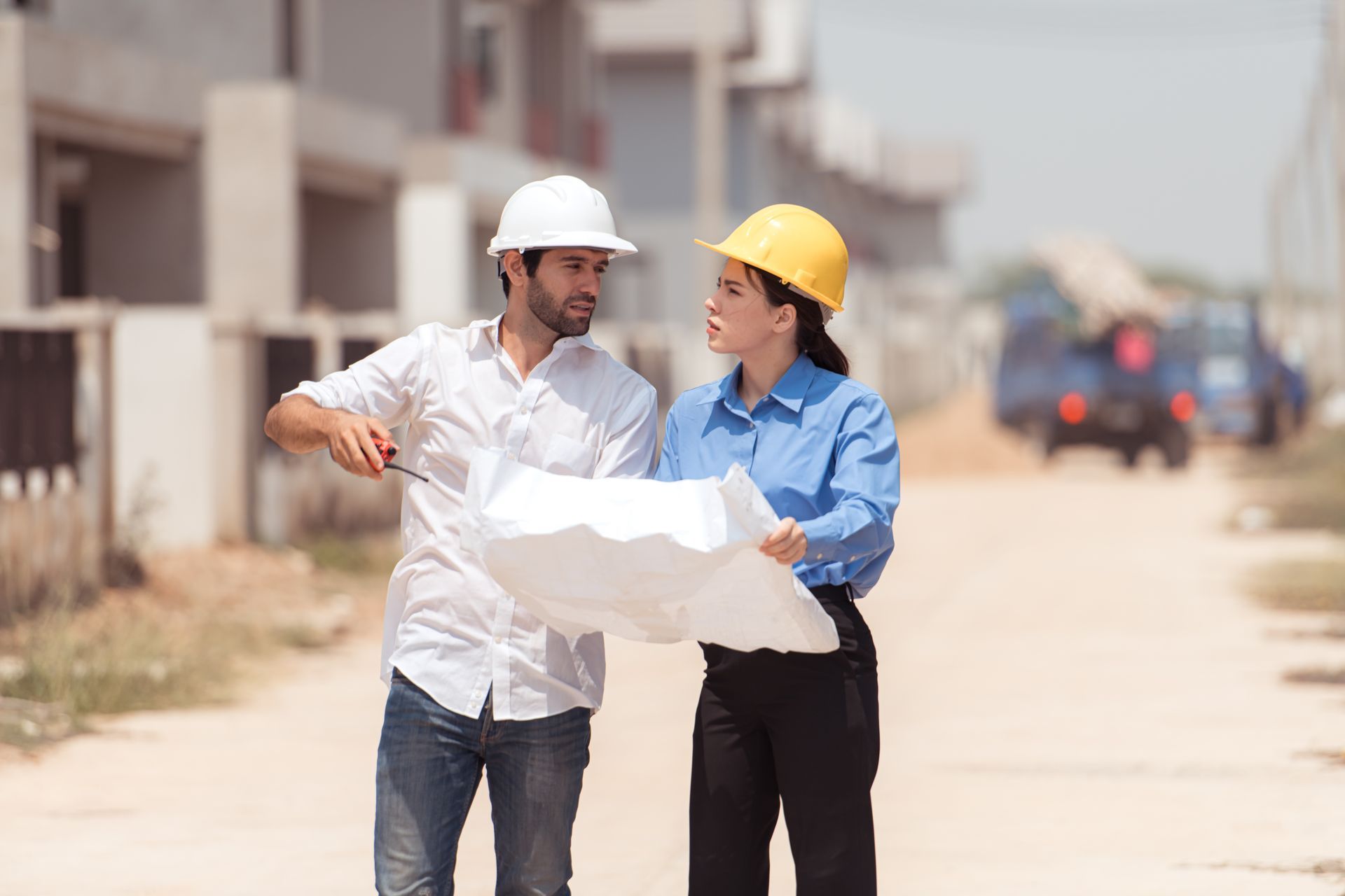 Two construction workers in hard hats examine blueprints outdoors.