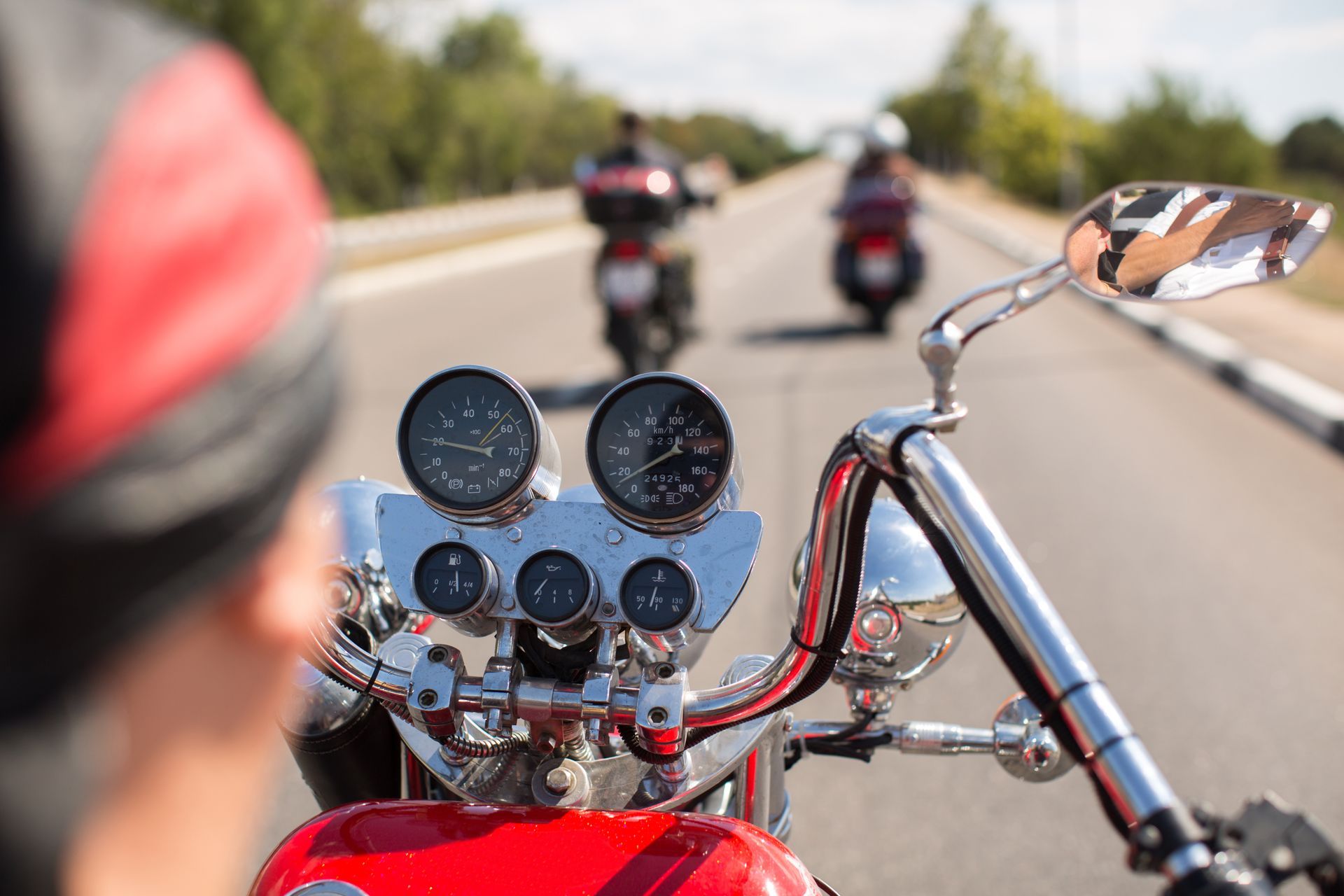 Motorcycle handlebars in focus, two motorcycles ahead on a sunny road, rider wearing red bandana.