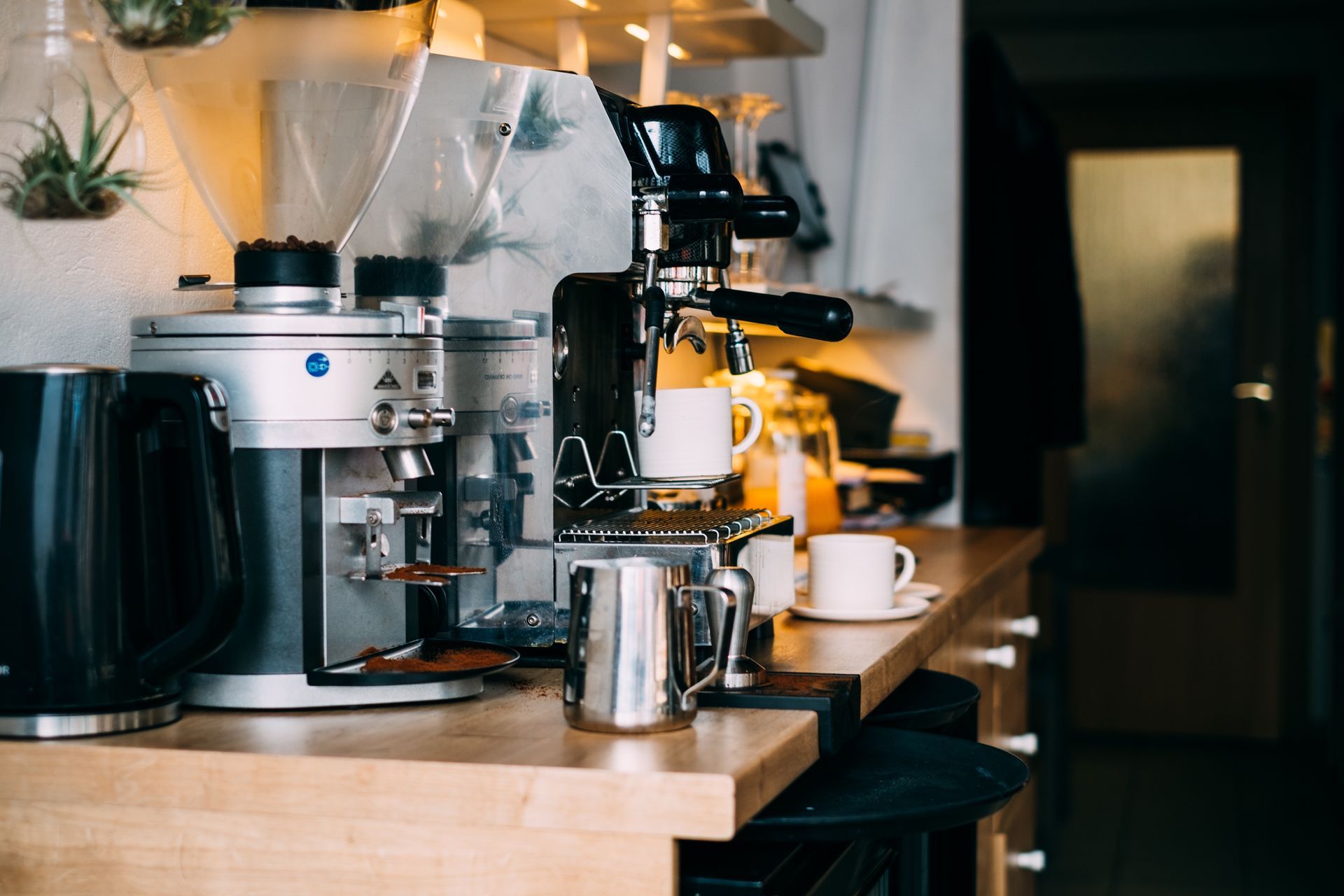Espresso machine and grinder on a wooden countertop, with cups, a kettle, and milk frothing pitcher in a kitchen.