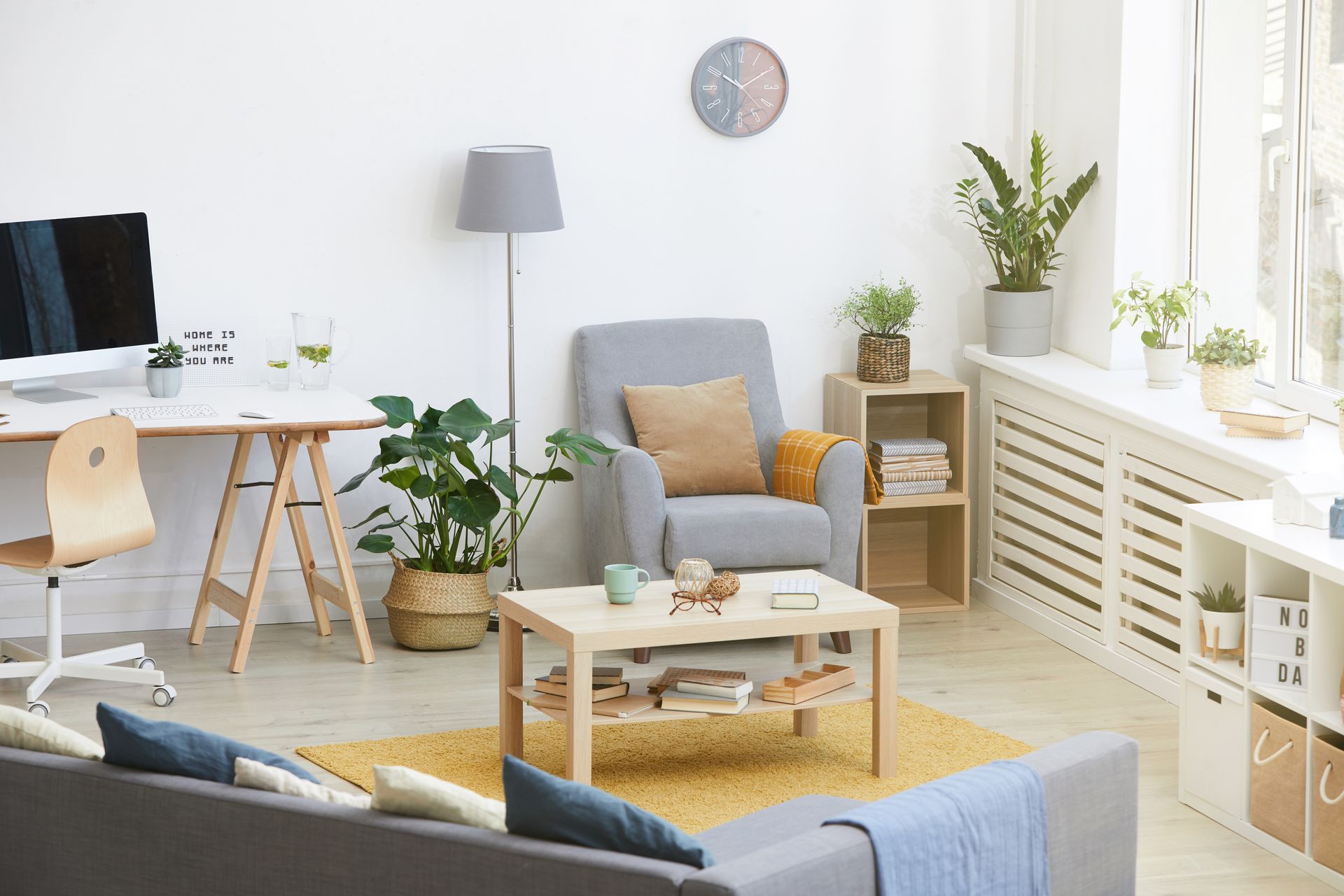Living room with gray sofa, armchair, desk, plants, wooden shelves, and yellow rug.