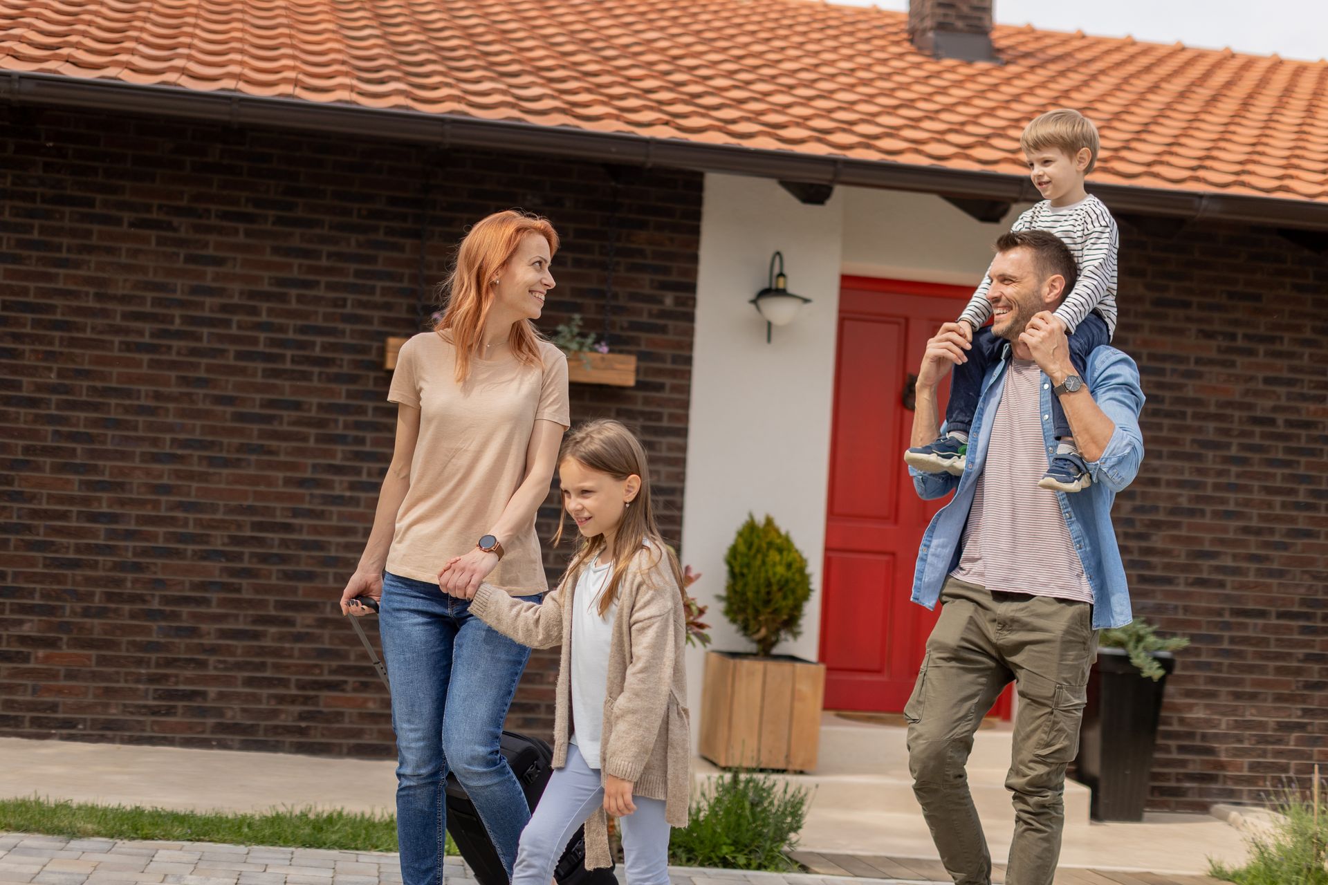 Family of four outside a house; mother holds hands with daughter, father carries son on shoulders.