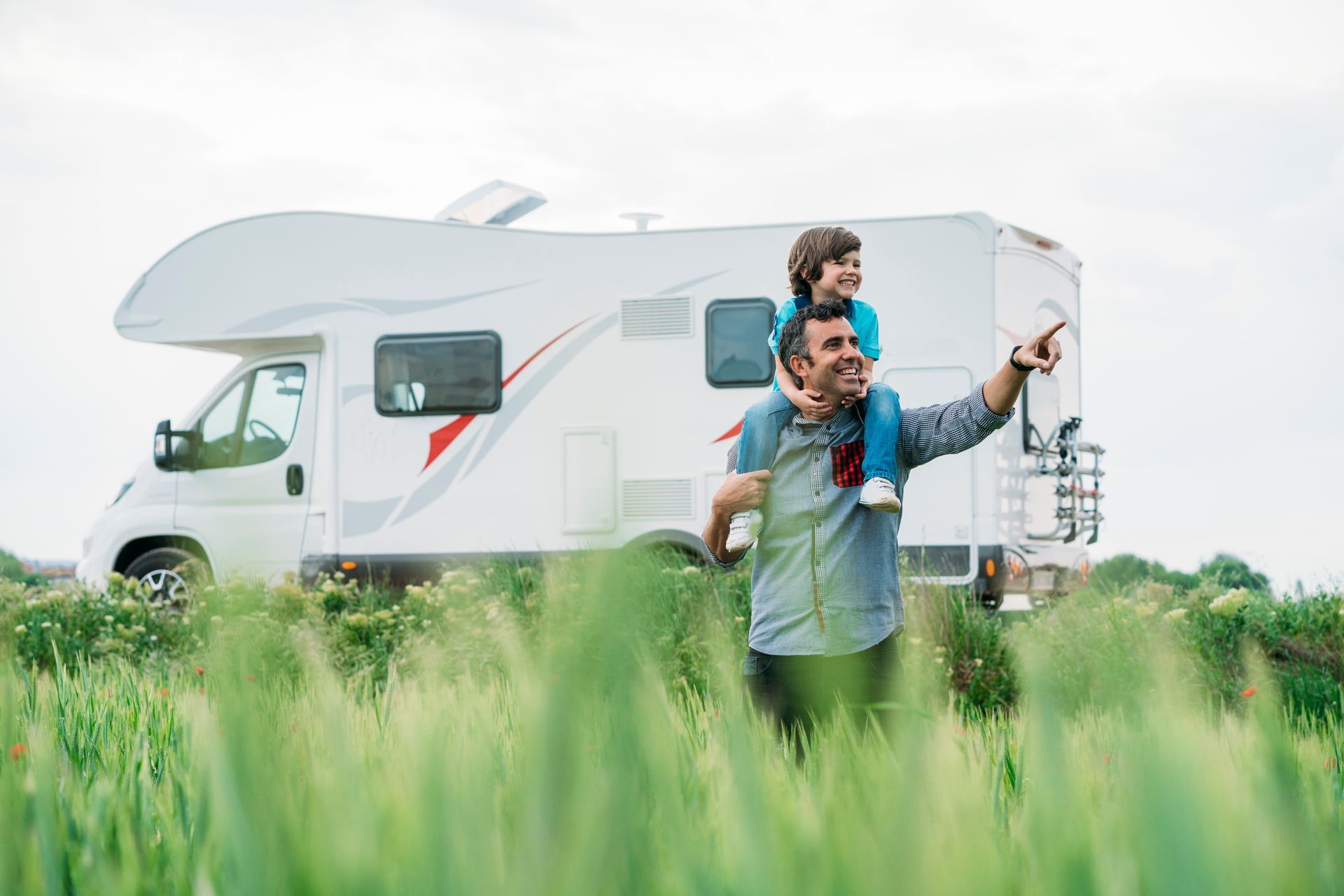 Man carrying child on shoulders points toward something, standing in tall grass next to a white RV.