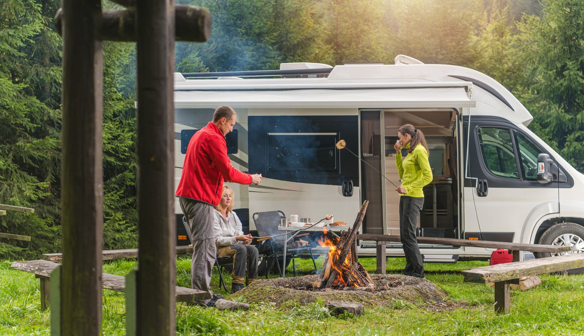 People camping by a campfire near a white RV in a grassy, wooded area.
