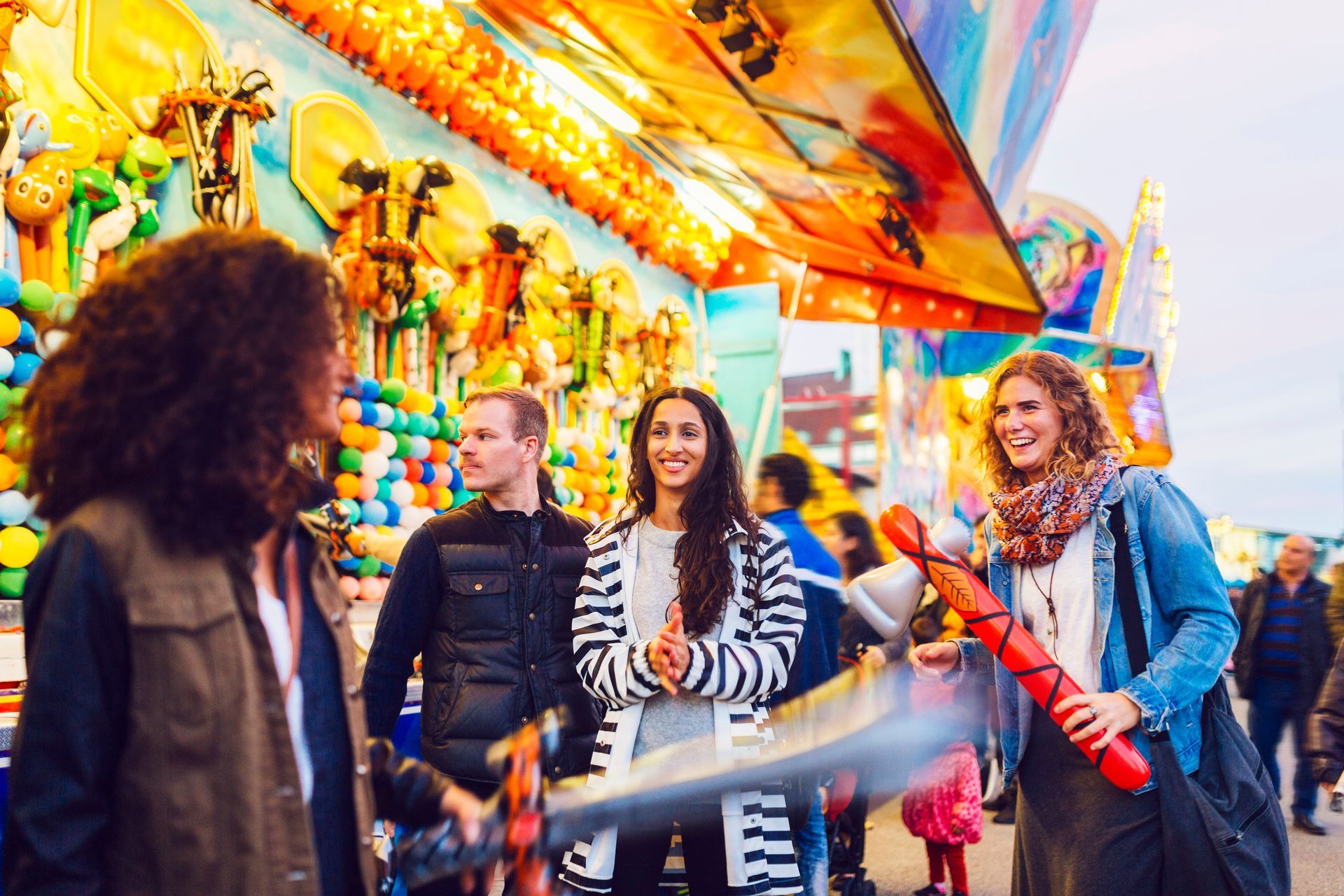 People at a carnival game, smiling and talking. Colorful lights and prizes in the background.