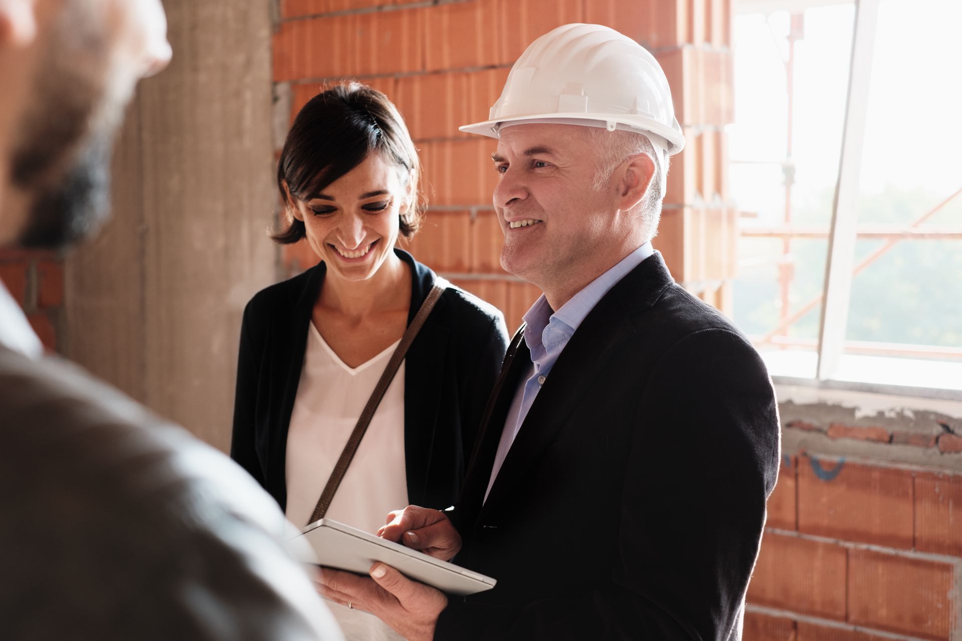 A construction foreman, in a hard hat and suit, smiles and discusses plans with two people in a building.