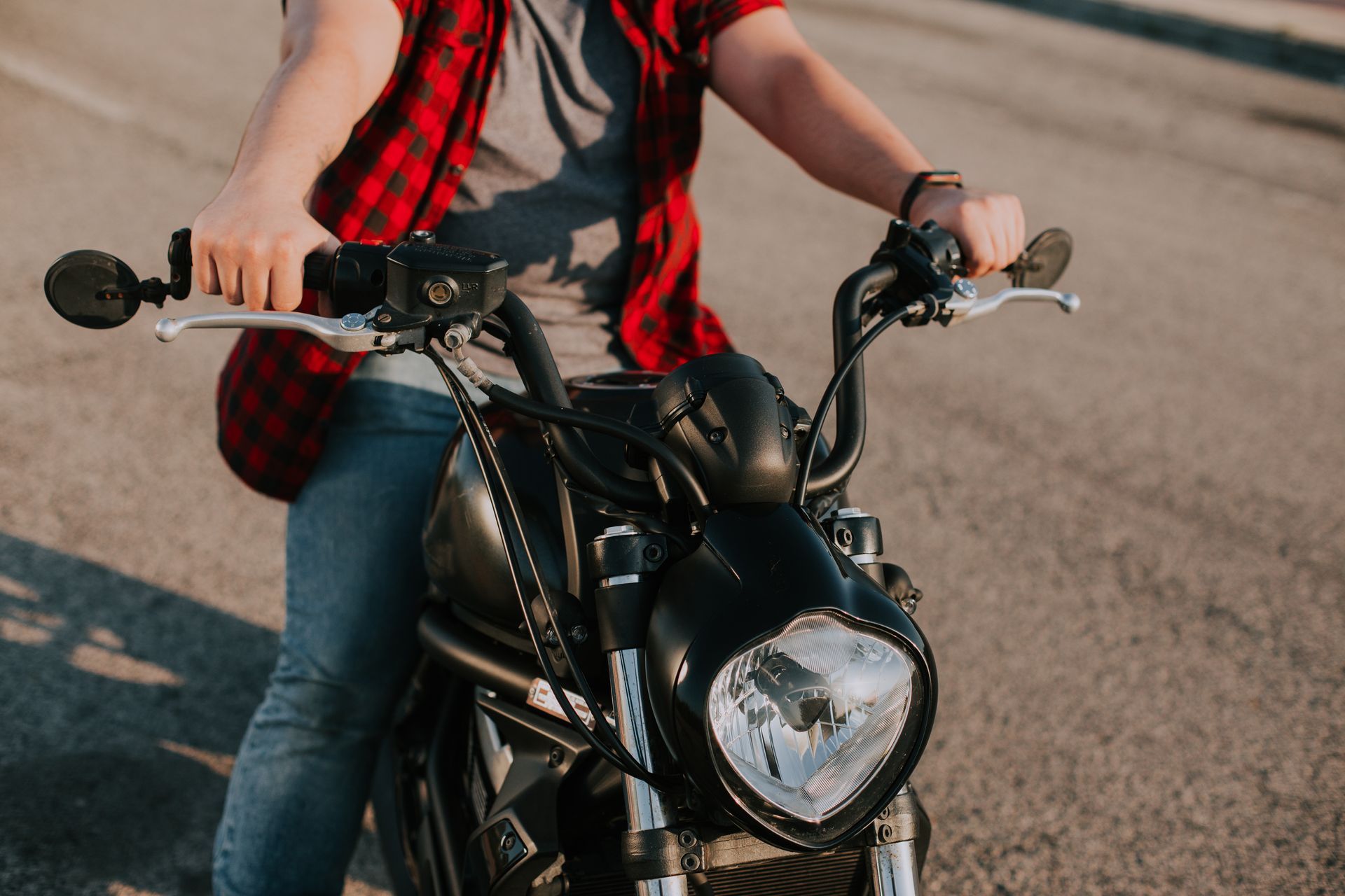 Person sitting on a black motorcycle on a road, wearing jeans and a red plaid shirt.