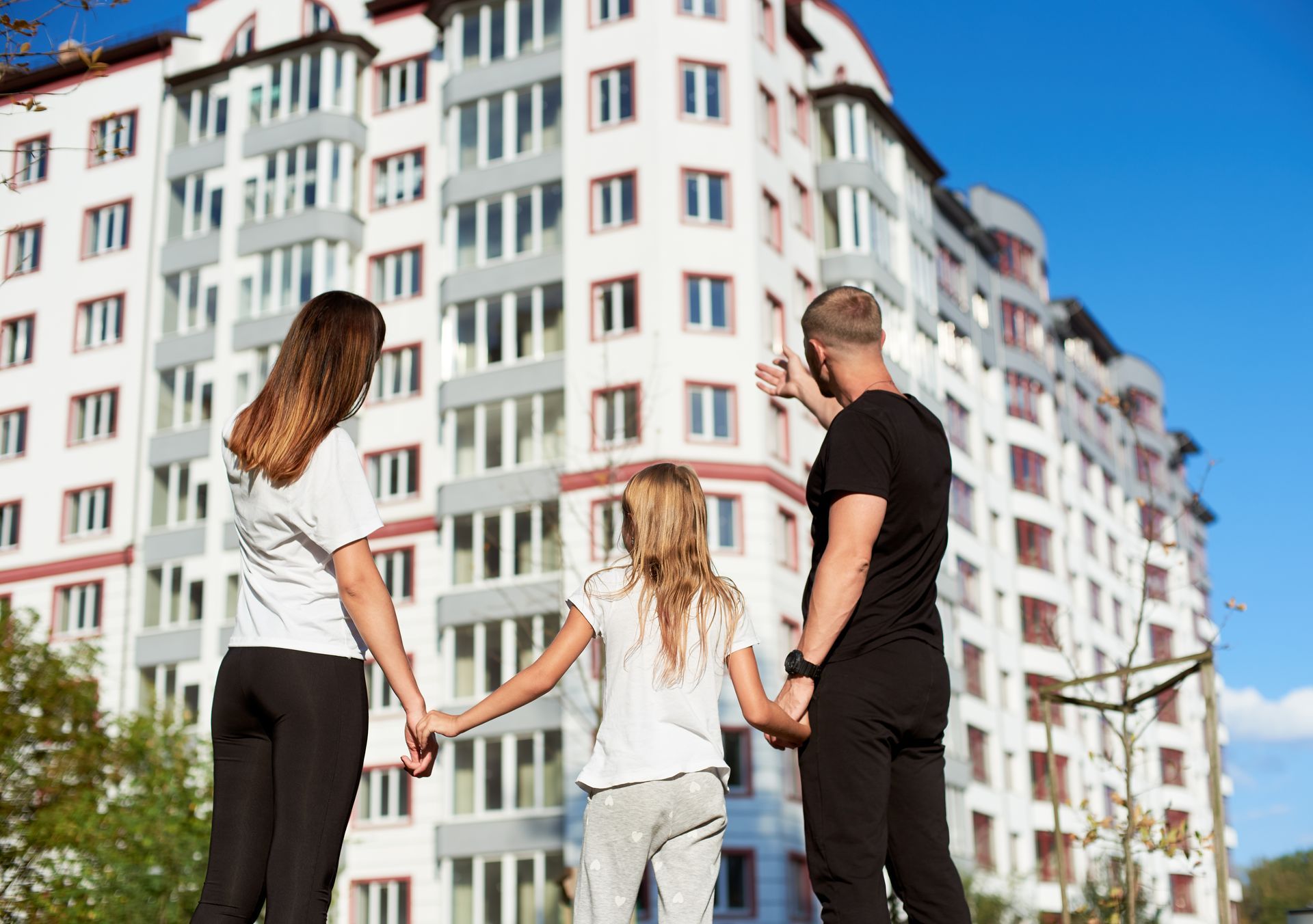 Family looking at a tall apartment building; man points upwards; sunny day.