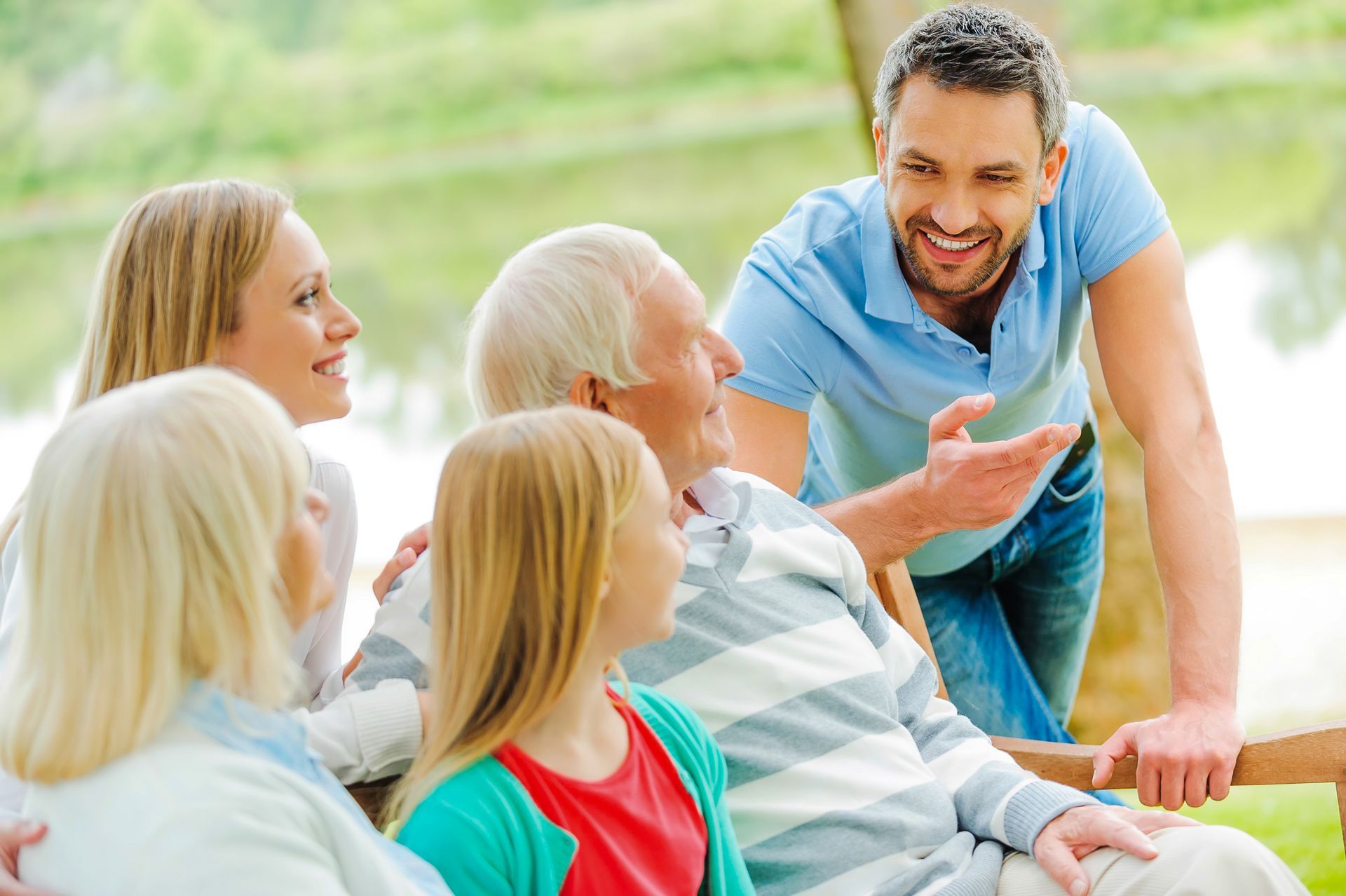 Family outdoors, smiling and talking; man gesturing, elderly man sitting, young girl, and blonde women.