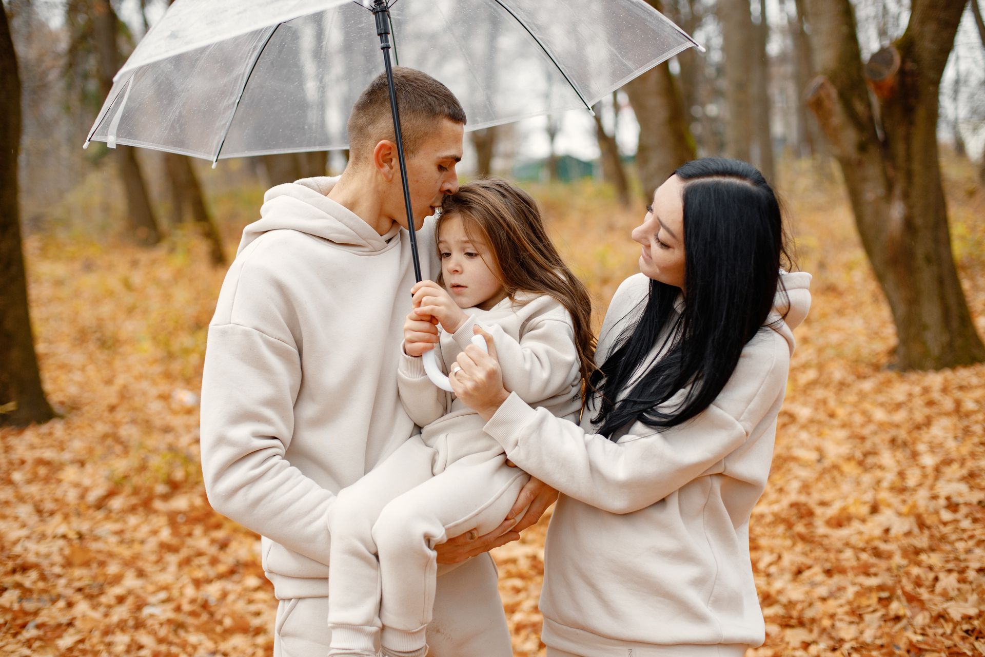 Family in matching beige hoodies under a clear umbrella in an autumn forest, embracing.