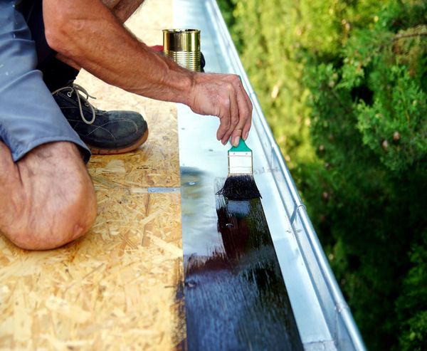 A man is painting a gutter on a roof with a brush.