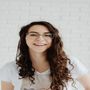 A woman wearing glasses and a white shirt is smiling in front of a white brick wall.