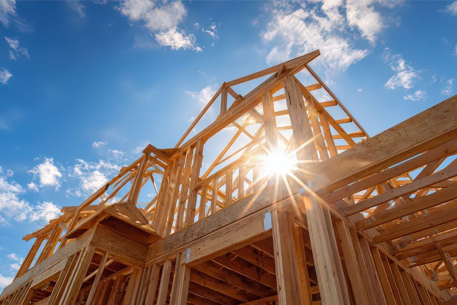 A low-angle view of a wooden house frame under construction against a bright blue sky with a sun flare.