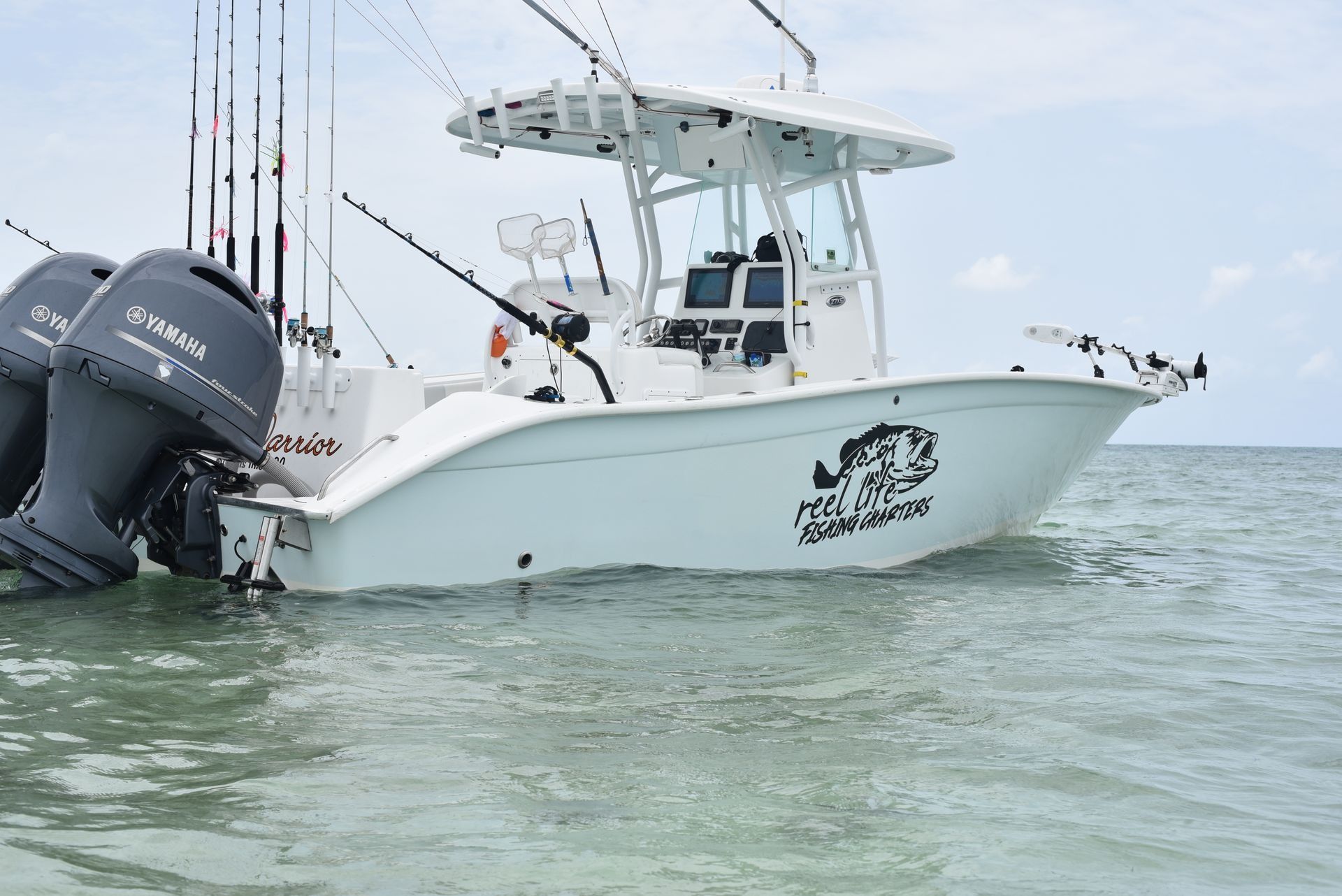 White fishing boat with several rods on calm water, twin outboard motors visible at the stern.