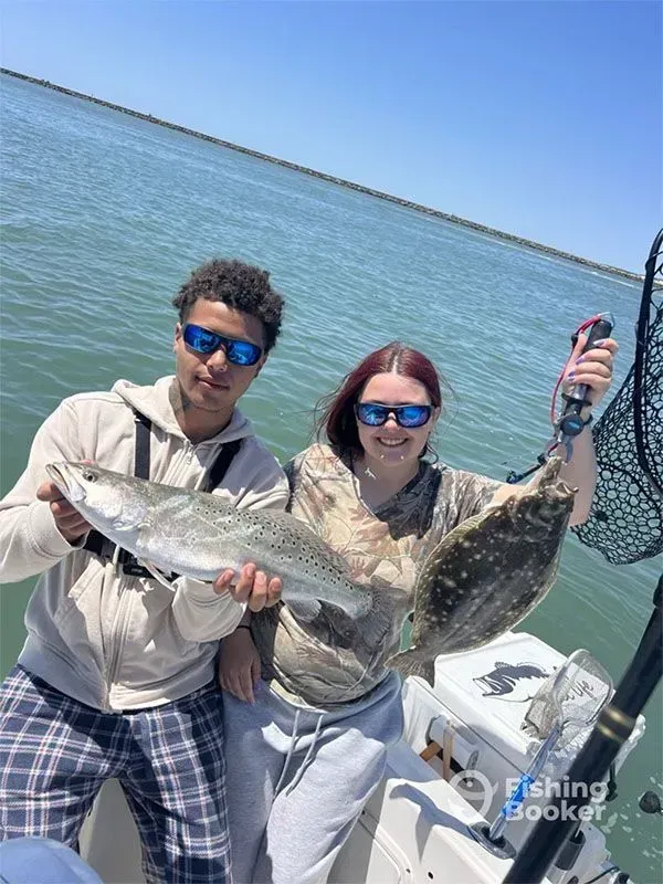 Two people on a boat holding large fish, smiling in sunglasses on calm blue water.