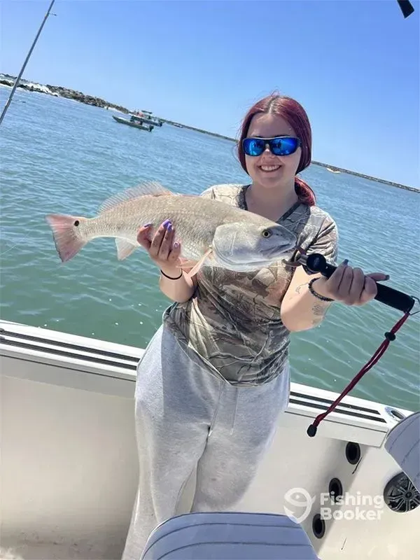 Person on a boat holding a large fish, wearing sunglasses, with water and shoreline in the background.