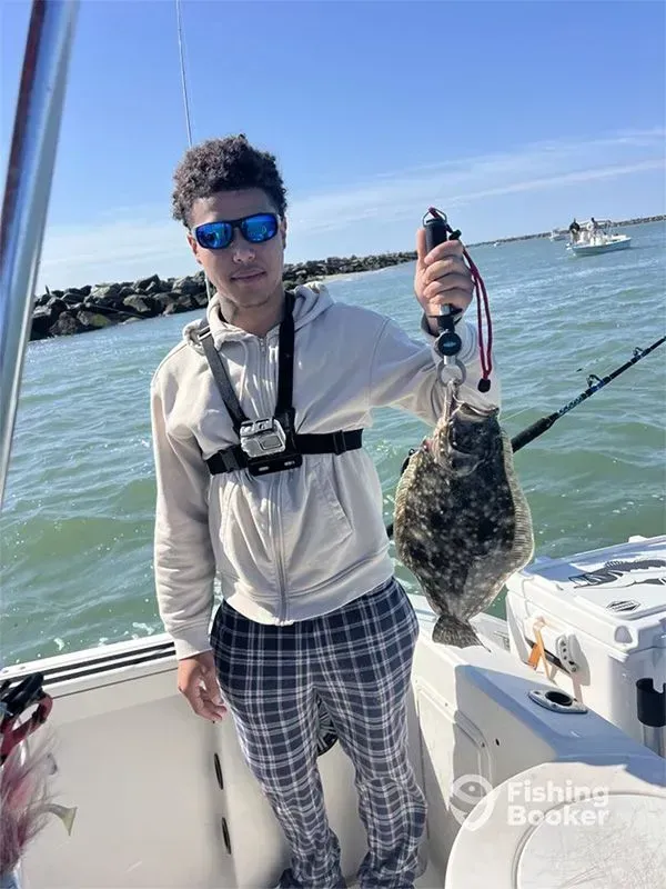 Person on a boat holding up a large fish, wearing sunglasses and plaid shorts on a sunny day.