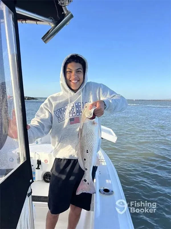 Person on a boat holding a fish over the water, wearing a white hoodie and black shorts.