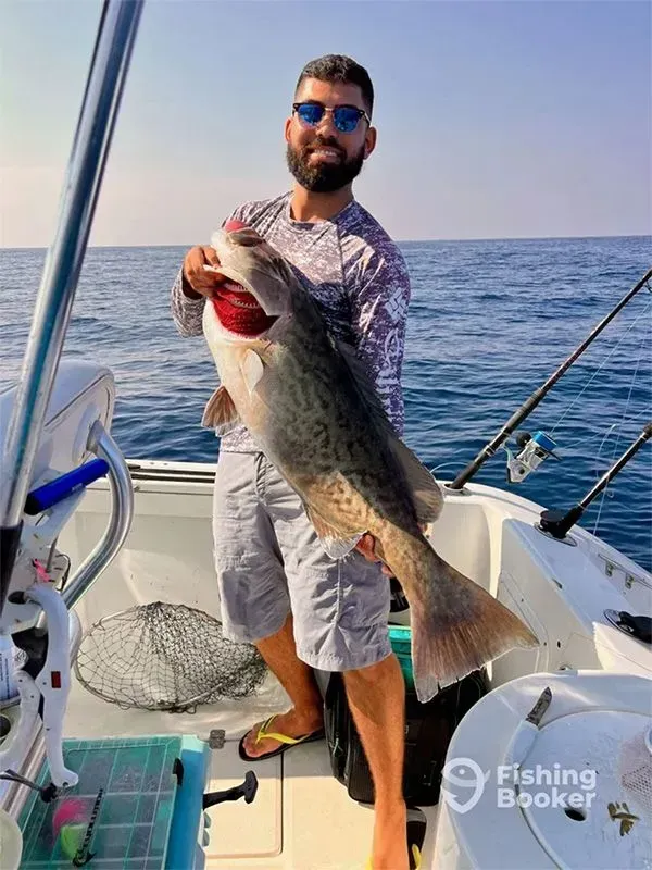 Man on a boat holding a large fish over the ocean, wearing sunglasses and a patterned shirt.