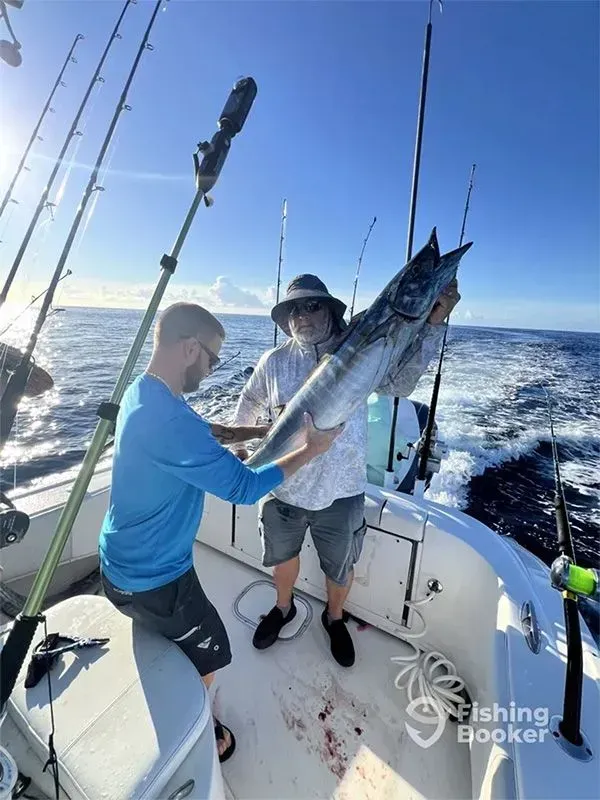 Two anglers on a boat hold up a large tuna at sea under a bright blue sky.