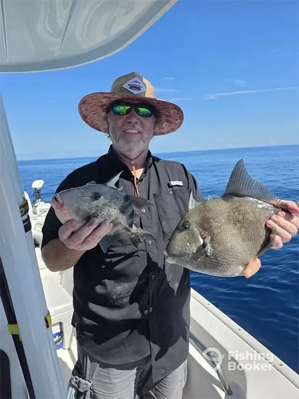 Man on a boat holding two fish, with blue ocean and sky behind him.