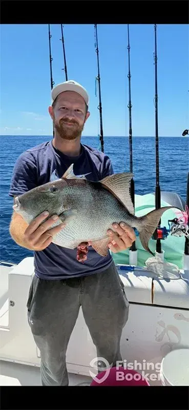 Man on a fishing boat holding a large fish, with ocean and fishing rods behind him.