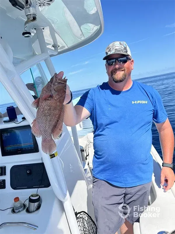 Man on a boat holding a large fish, wearing a blue shirt and camo cap under a sunny sky
