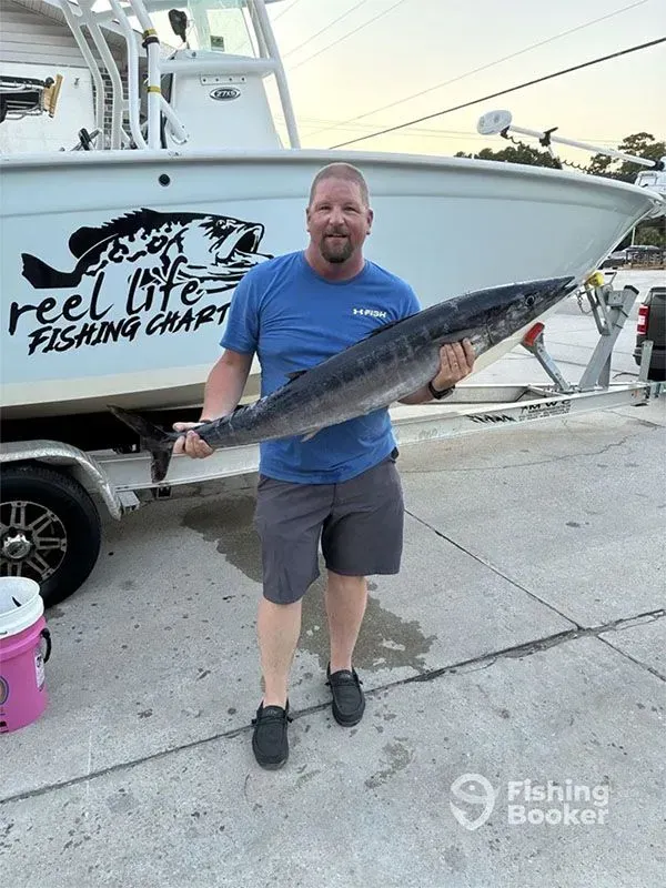 Man holding a large fish in front of a boat with “Reel Life Fishing Charters” logo at sunset