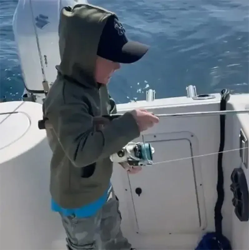 Child fishing from a boat, wearing a hooded jacket and cap, holding a rod over calm blue water