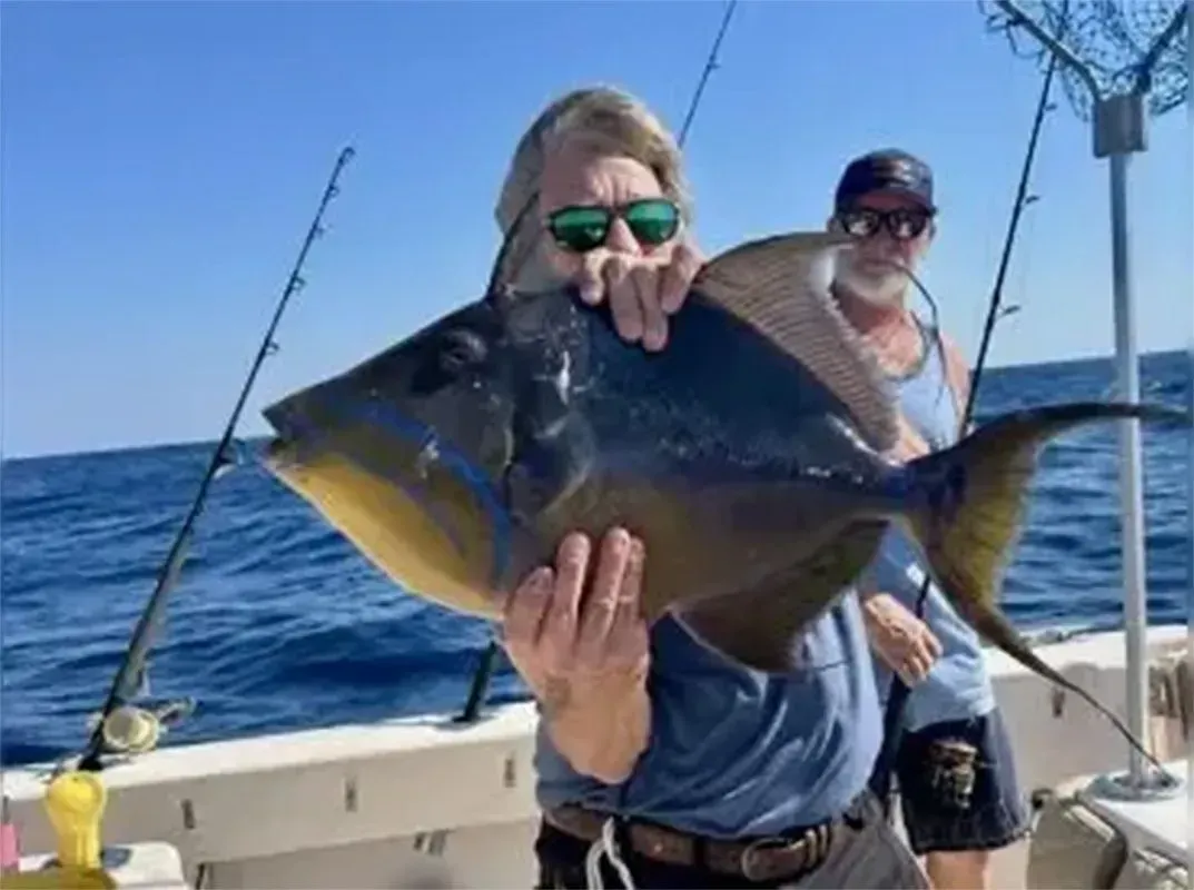 Two anglers on a boat holding a large dark fish in bright blue ocean waters