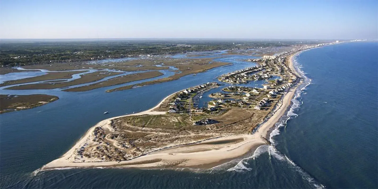 Aerial view of a narrow sandy barrier island with marshes on one side and open ocean on the other.