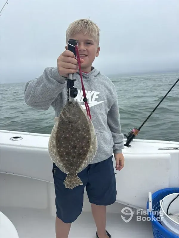 Boy holding up a flat fish on a boat, with fishing rod and calm water in the background.
