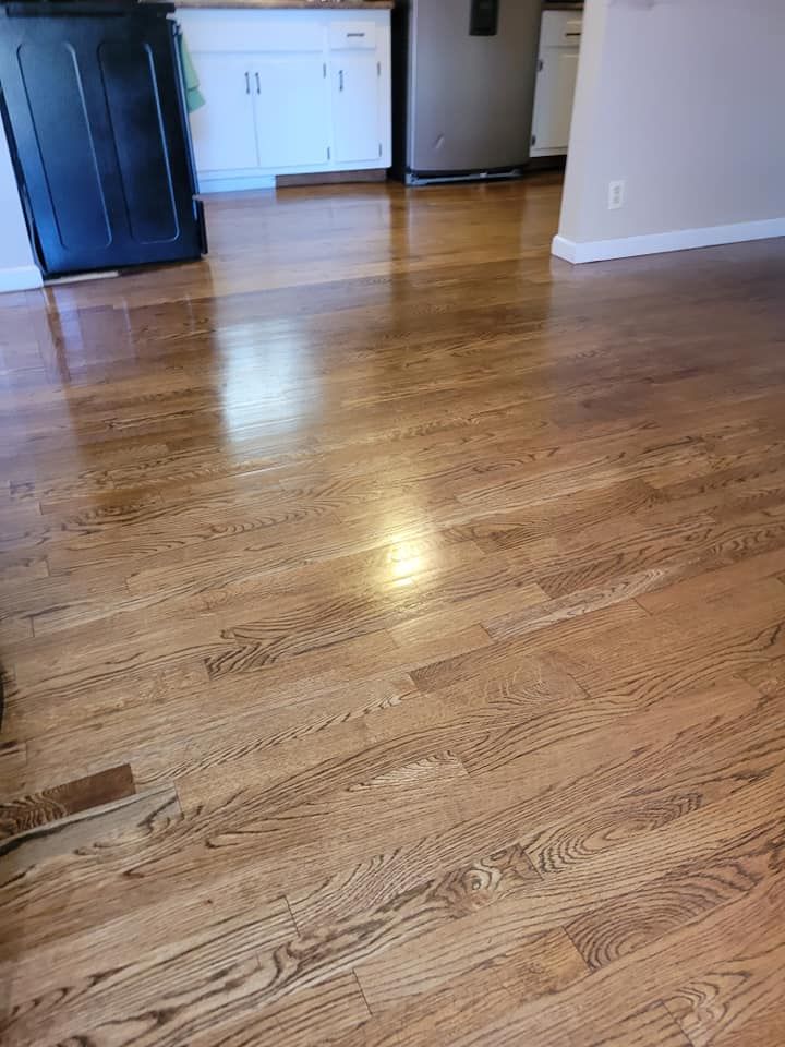 Polished, light-brown hardwood floor in a home, reflecting natural light. Kitchen cabinets are in the background.