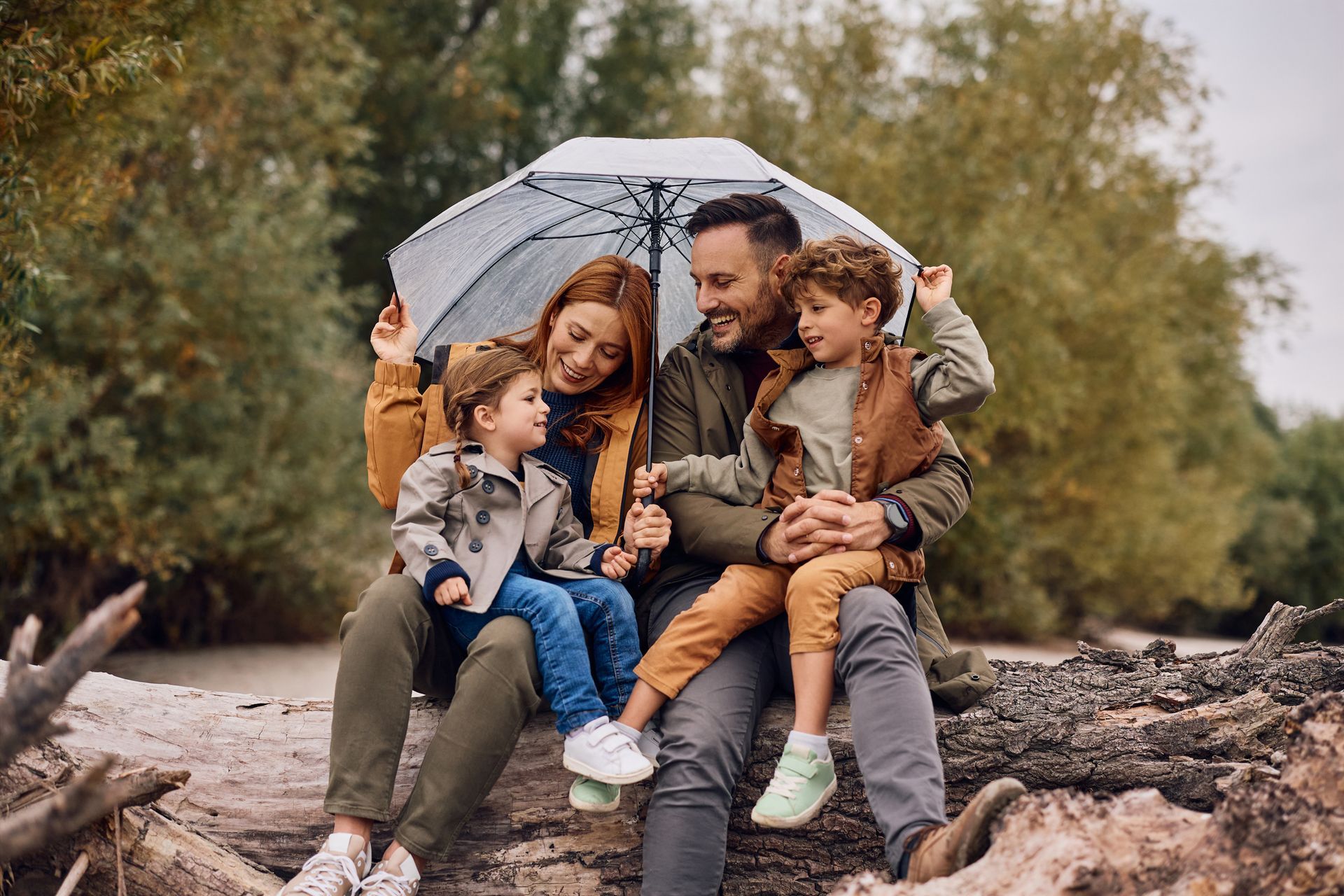 Family of four sits under umbrella, smiling, outdoors in autumn.