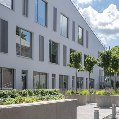 Modern gray apartment building with large windows, trees, and landscaped courtyard under a bright sky