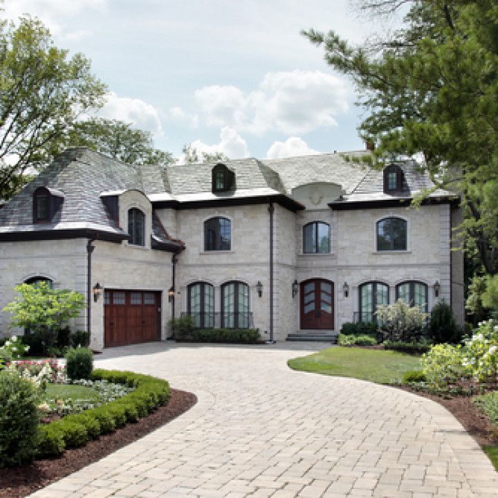 Large gray stone mansion with a curved driveway, landscaped garden, and trees