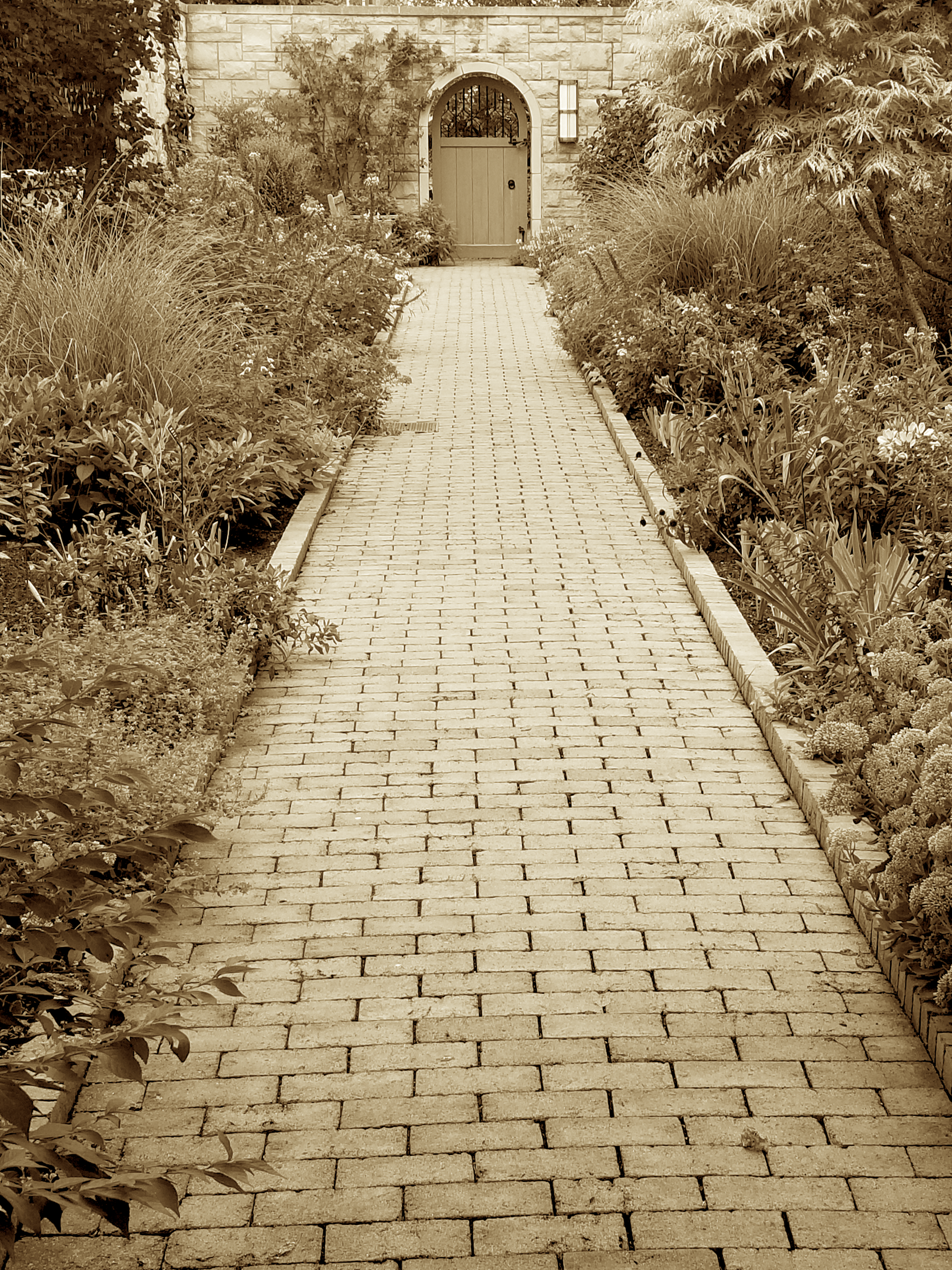 Brick pathway leading to a doorway in a stone wall, flanked by bushes and plants. Sepia tone.