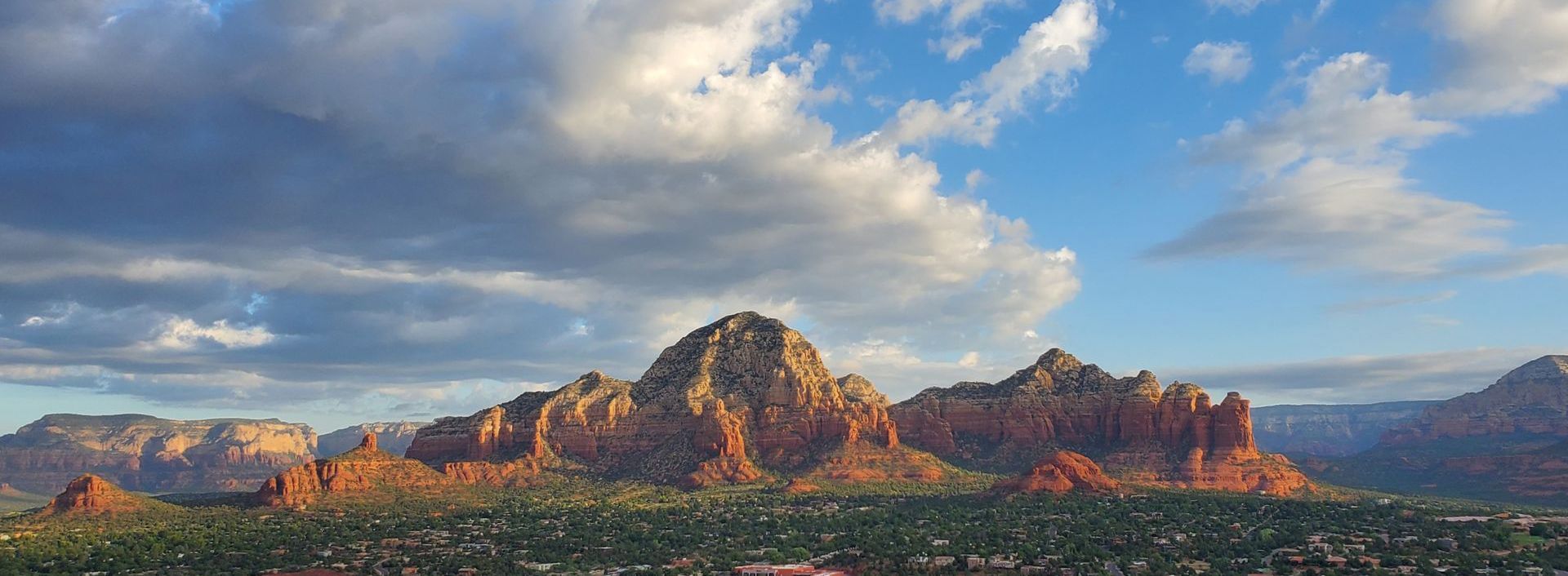 Red rock formations and green trees under a partly cloudy sky.