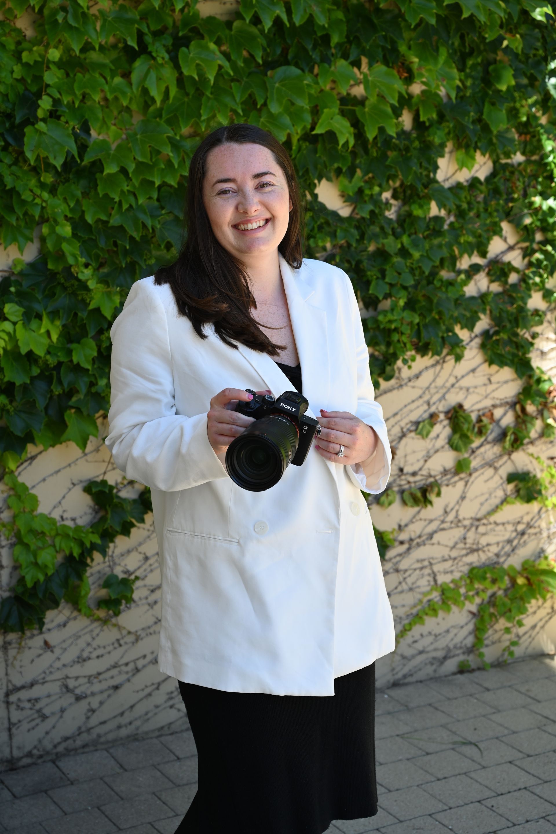 Woman in white jacket holding a camera, smiling in front of ivy-covered wall.
