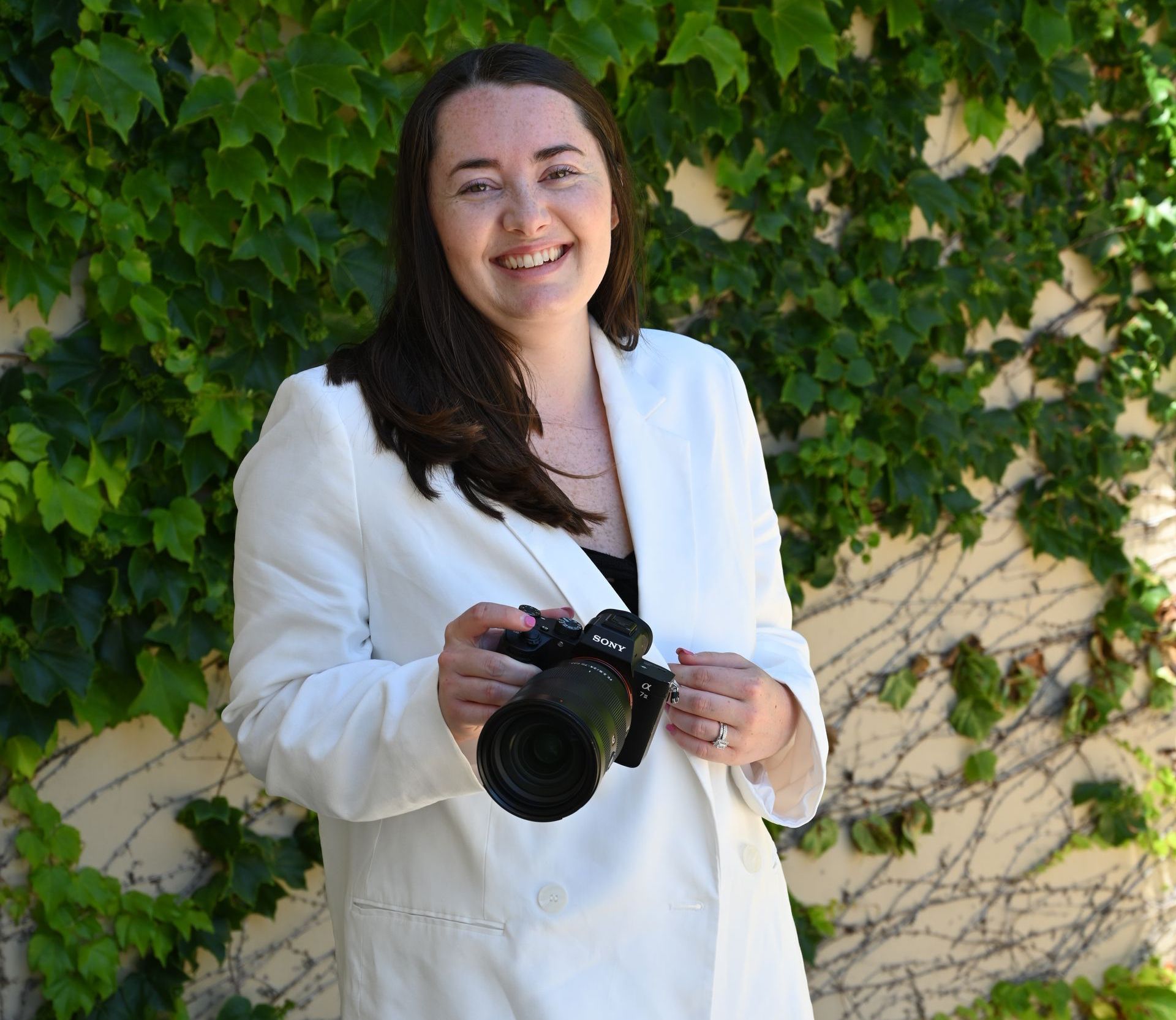 Woman in white blazer holds a camera, smiles, stands in front of a green ivy wall.