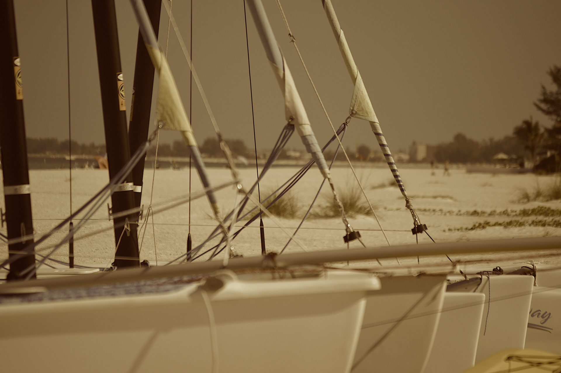 Sailboats lined up on a sandy beach, masts and rigging visible, muted tones.