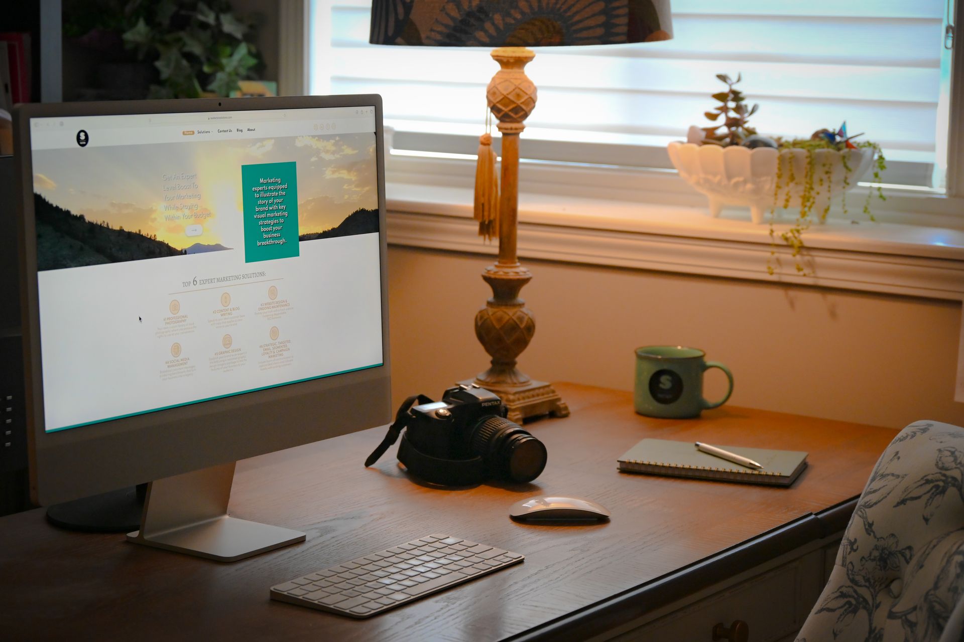 Modern desk with computer monitor, keyboard, lamp, camera, and window in a warm home office