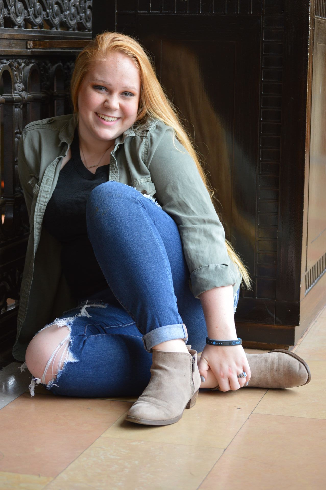 Woman with long red hair, smiling, wearing olive green jacket, ripped blue jeans, sitting, light brown ankle boots.