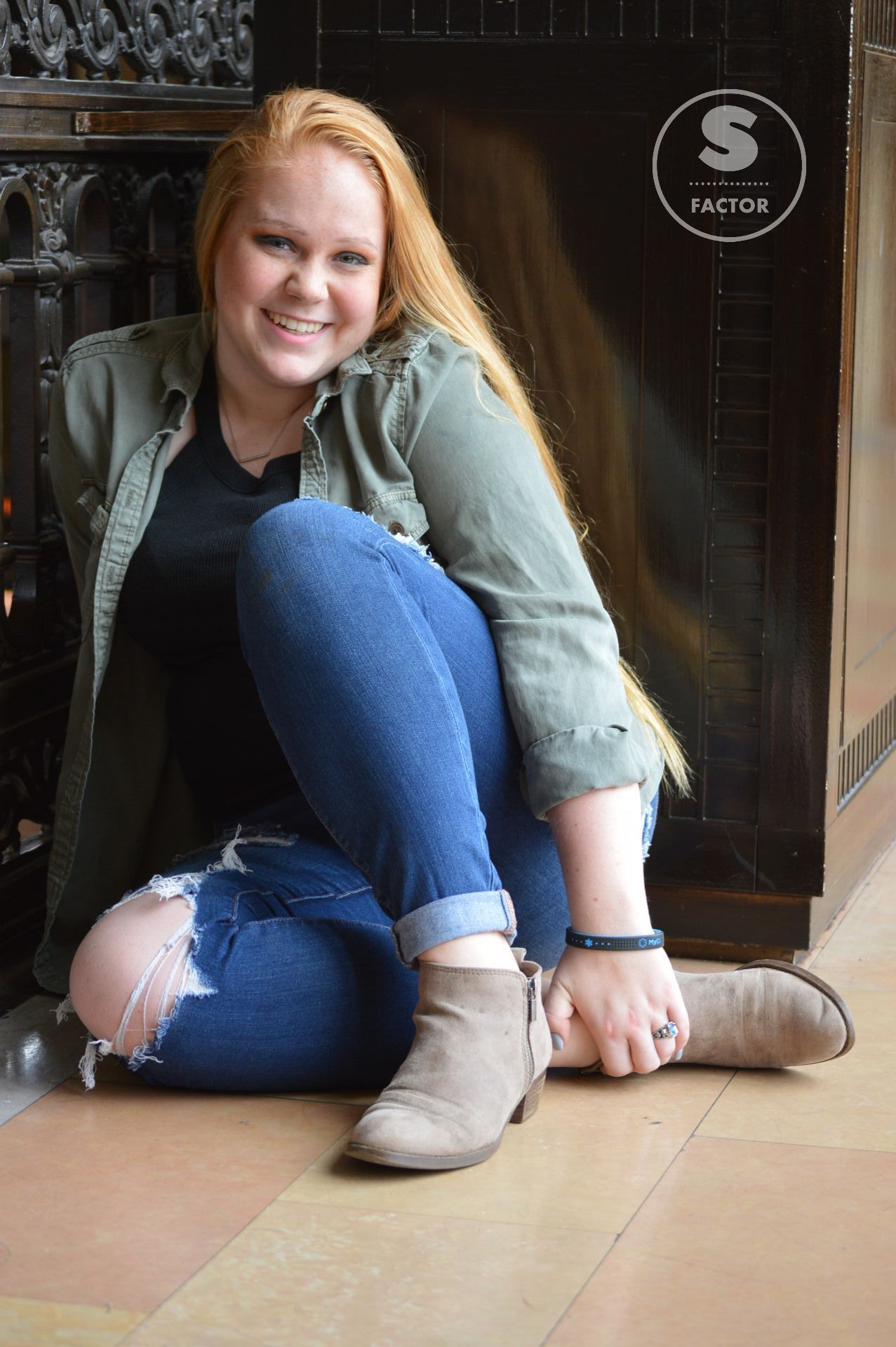 Woman with long red hair, smiling, in jeans, jacket, and boots, posed indoors.