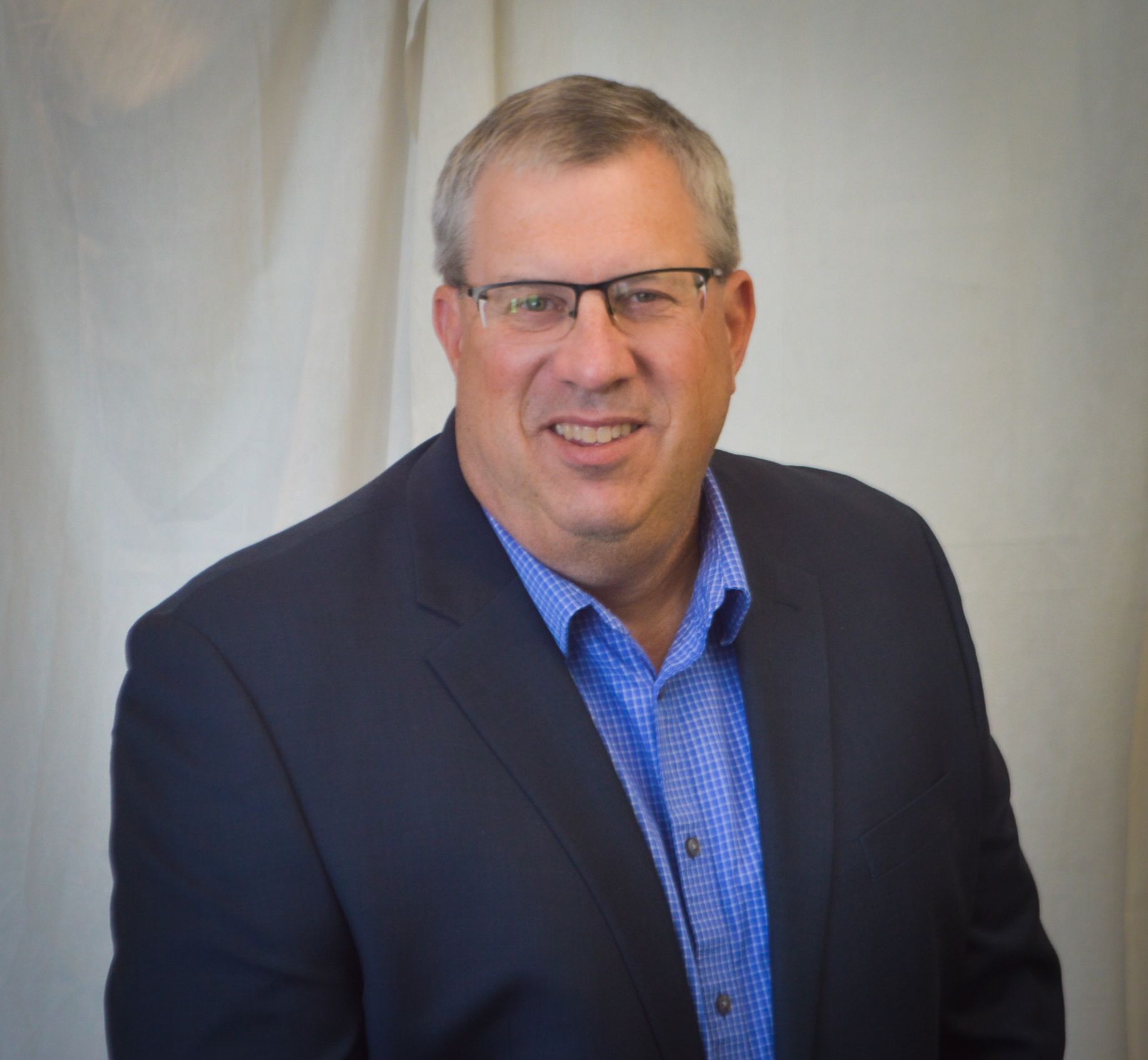 Man in glasses, blue shirt, and navy blazer, smiling. White backdrop.