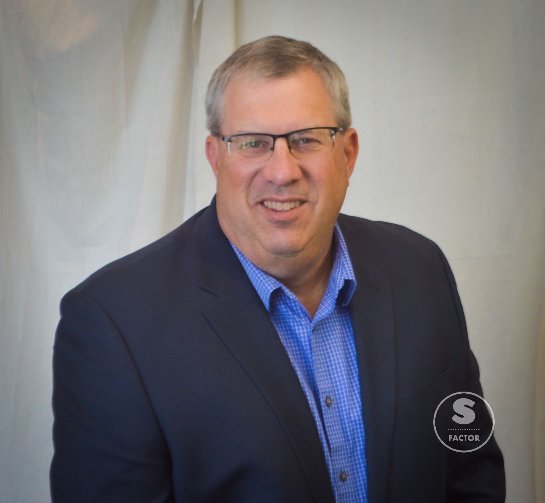 Man in glasses, blue button-down shirt, and navy blazer smiles against a white backdrop.