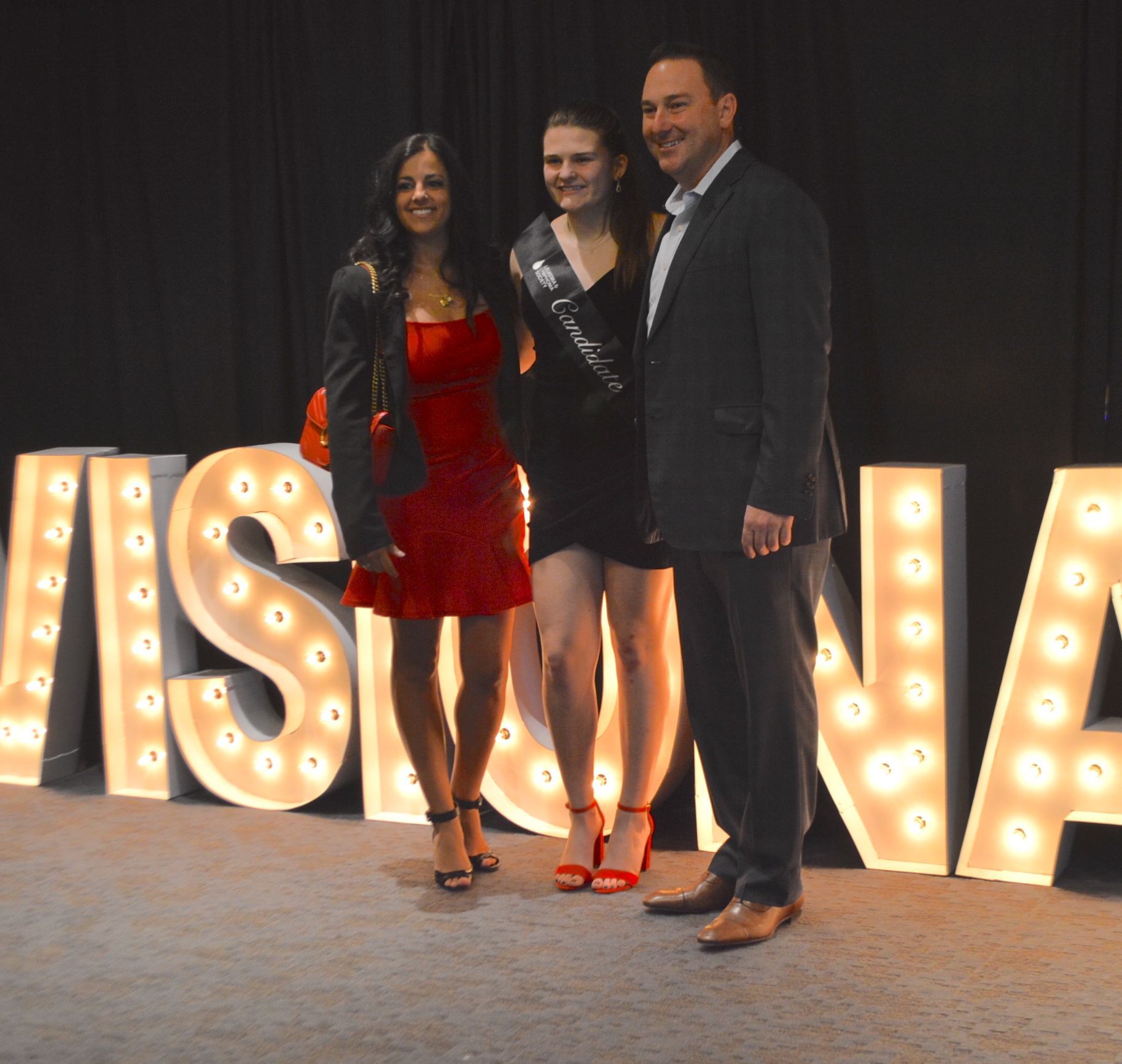Three people pose in front of glowing letters. Woman in red dress, woman in black dress with sash, man in suit smile.