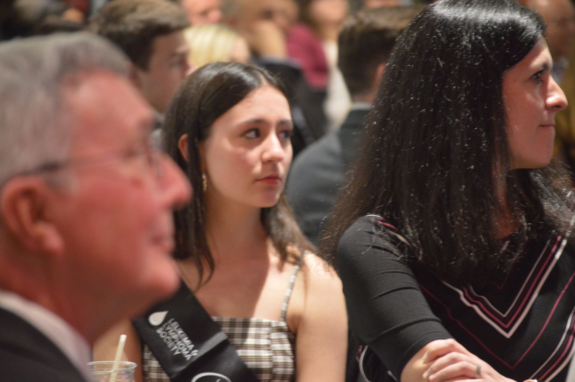 People seated indoors, listening attentively. A woman in a checkered dress and another with dark hair are visible.