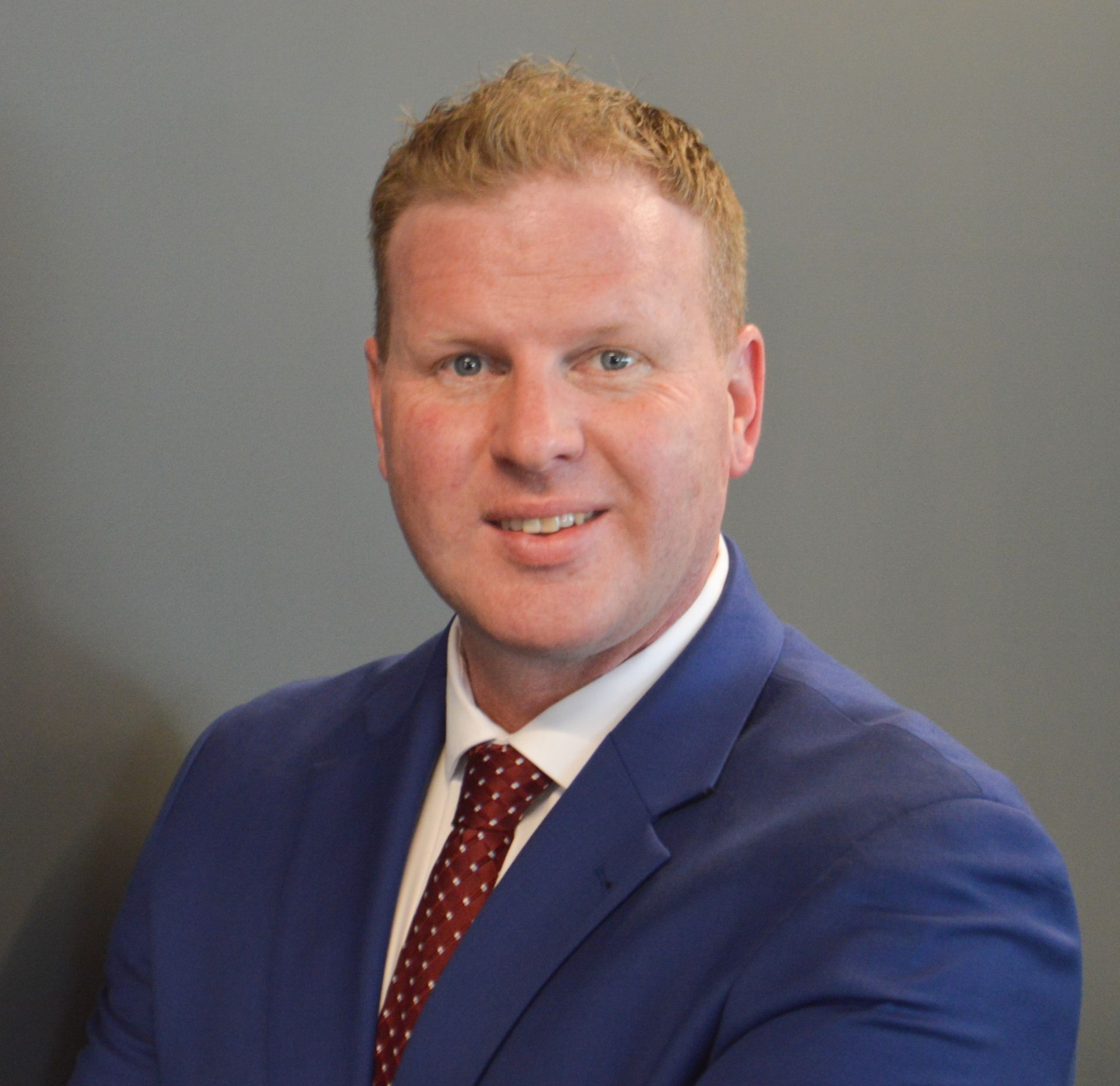 Man in blue suit, red tie, smiling in front of gray background.