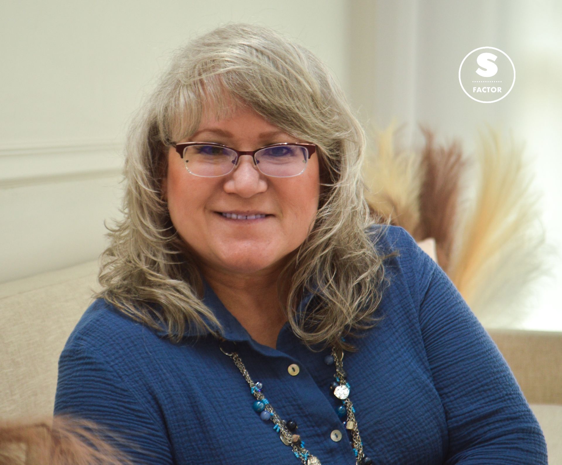Woman with glasses and a blue shirt smiles. She sits indoors with neutral-colored decor behind her.