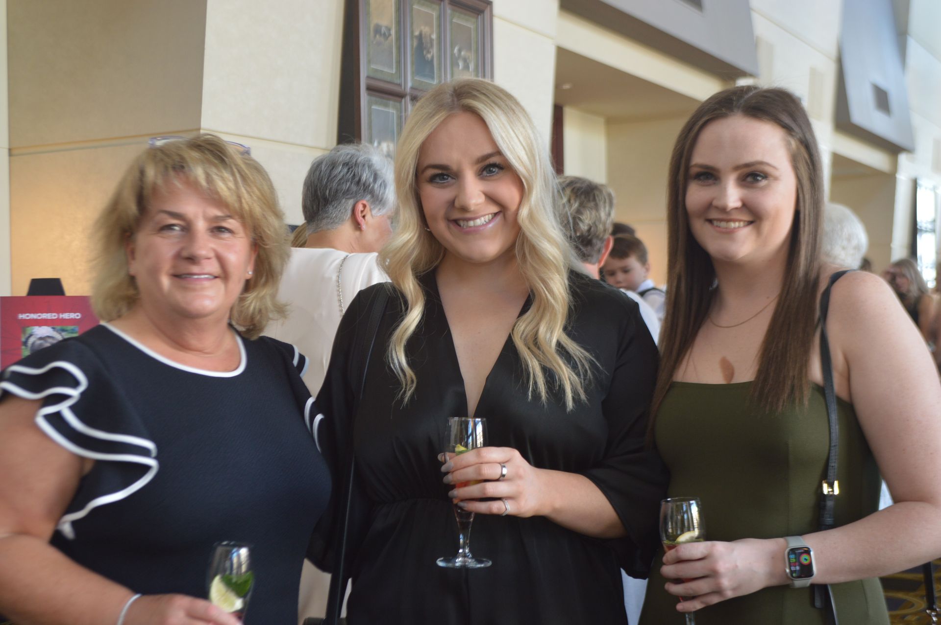 Three women smiling, holding drinks, standing indoors. One in black dress, two in green and navy tops.